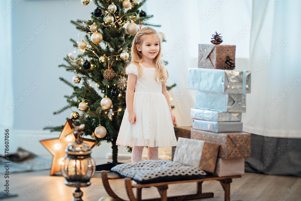 a beautiful little girl in a white dress puts gifts on a sled near the Christmas tree. happy new year. retro-style. interior decoration for a holiday.