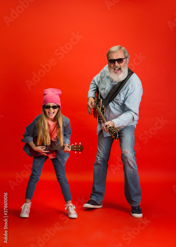 Music band. Senior man spending happy time with granddaughter on red background. Joyful elderly lifestyle, family, childhood, tech concept. Enjoying modern music, technologies. Copyspace. photo