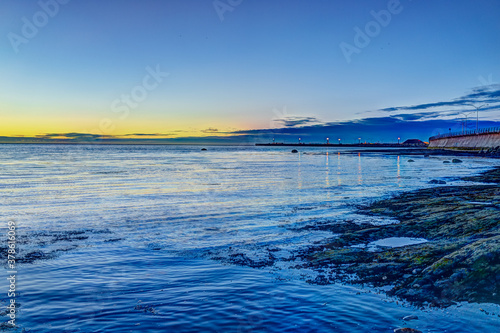 Yellow orange sunset twilight in Rimouski, Quebec by Saint Lawrence river in Gaspesie region of Canada with rock boulders in shallow water with pier photo