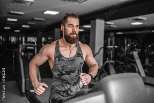 Adult strong bearded man running on treadmill in gym.