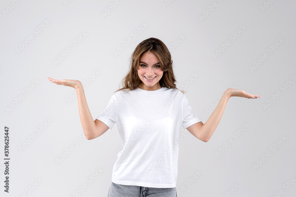 Smiling young woman standing against white background and holding her hands up as scales for invisible objects, looking to the camera Smiling young woman standing against white background and holding her hands up as scales for invisible objects, looking to the camera