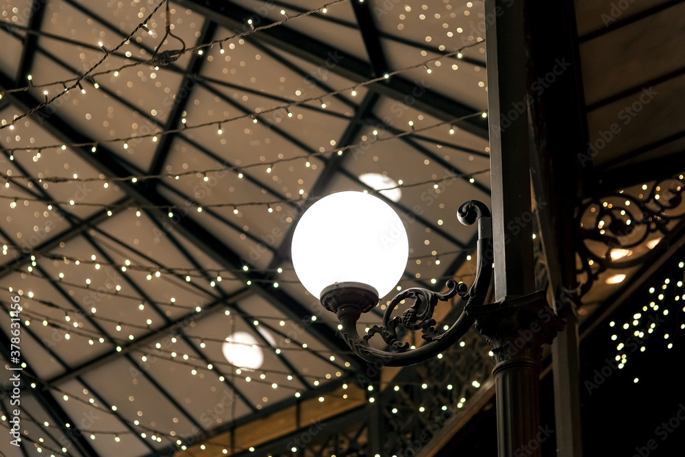 Fototapeta premium white round lantern on a wrought iron pillar of the restaurant terrace gazebo with garlands on the ceiling, architectural lighting of the room in a retro style, nobody.