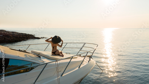 Attractive woman with big hat sitting at white luxury yacht with sea and sun track on water at background