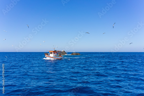 Orange and white fishing boat followed by seagulls on aegean sea. blue sky and sunny day