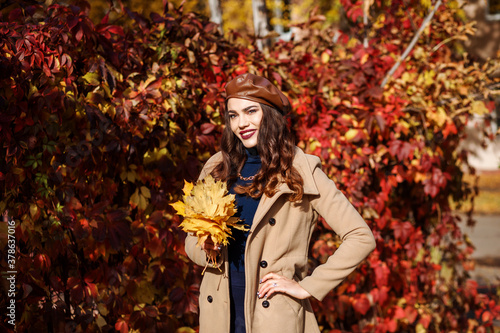Fashionable attractive woman in front of the red leaves hedge outdoors in autumn. Stylish woman is wearing brown leather beret and beige coat, dark blue dress. Yellow maple leave in the model's hands.
