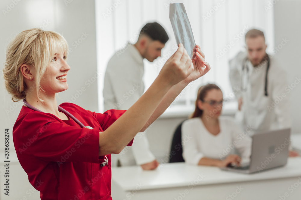 Obraz premium Young female doctor in hospital looking at x-ray. The other two doctors communicate in the background. Blonde doctor in the office with an X-ray. Medicine and treatment concept