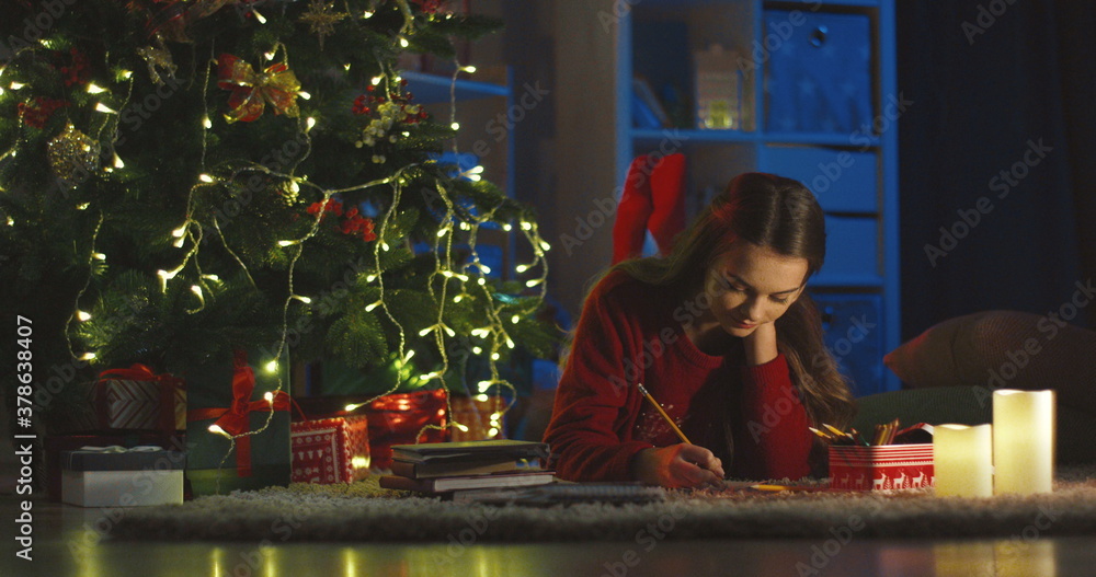 Caucasian teenage pretty girl lying on the floor in the room decorated for the Christmas with a tree and drawing a picture or writing a x-mas postcard with greeting.