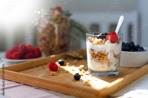 muesli yogurt with berries standing on a bamboo wooden tray in sunlight. breakfast muesli jar in the background. blueberries and rapsberries as decoration.