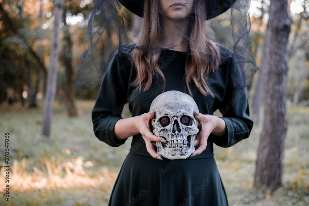 Young beautiful cute woman in dark dress and witch's hat holds a skull in her hands. Halloween party costume. Forest, park with autumn trees. Young beautiful cute woman in dark dress and witch's hat holds a skull in her hands. Halloween party costume. Forest, park with autumn trees.