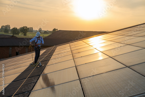 Maintenance assistance technical worker in uniform is checking an operation and efficiency performance of photovoltaic solar panels on roof at sunset.