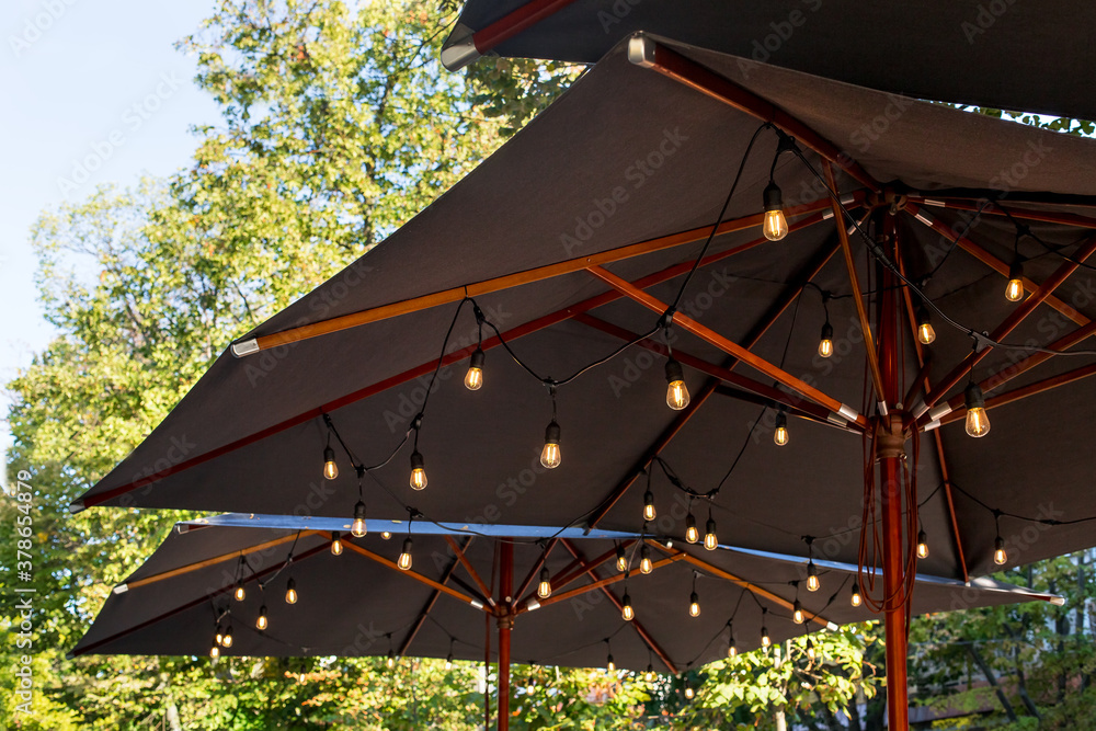textile umbrella with a wooden frame and edison pendant lamps glowing with warm light on a backyard terrace against a background of green trees with a sky, nobody.