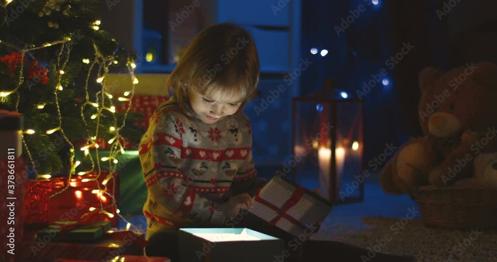 Portrait of the pretty small Caucasian girl opening a present x-mas box and looking inside as it shining on Christmas night. Portrait of the pretty small Caucasian girl opening a present x-mas box and looking inside as it shining on Christmas night.