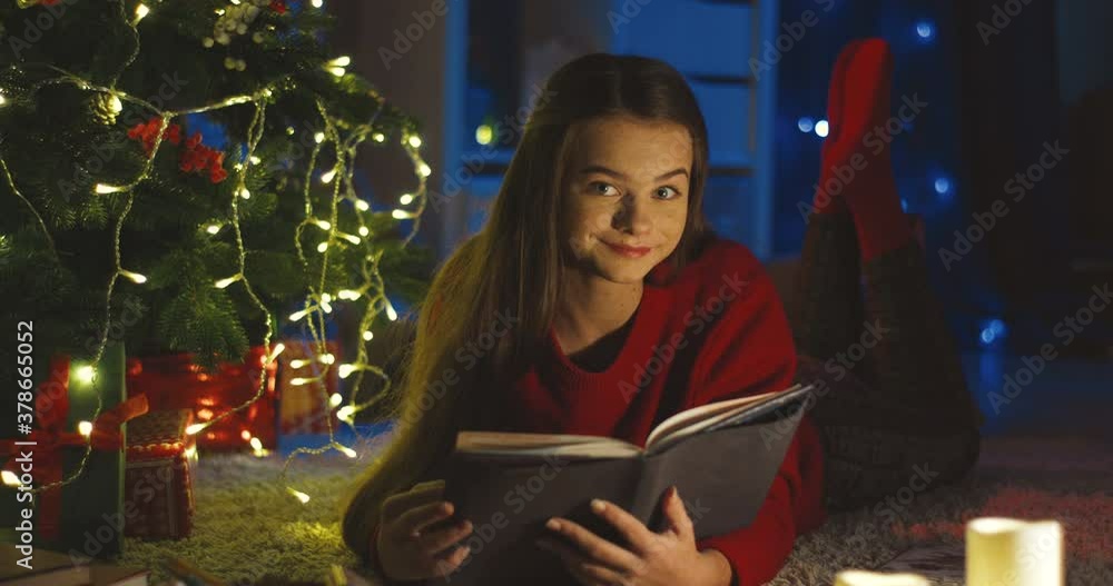 Portrait of the beautiful teenager girl lying under the Chritmas tree and reading a book, then smiling to the camera. Portrait of the beautiful teenager girl lying under the Chritmas tree and reading a book, then smiling to the camera.