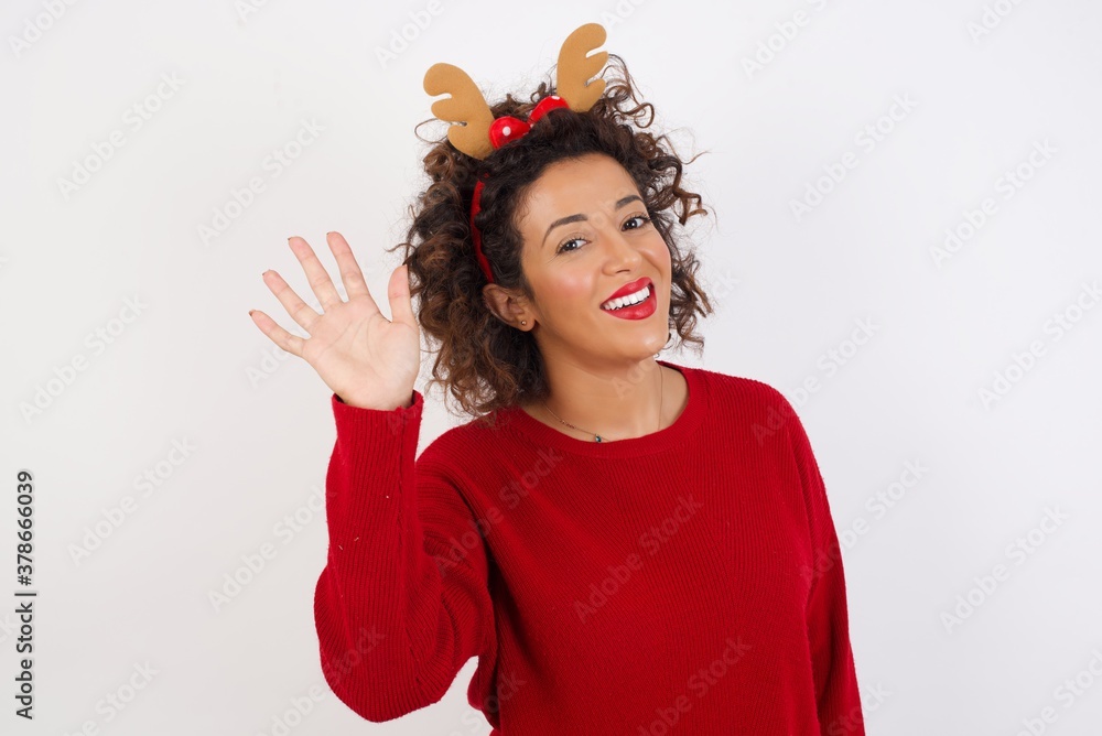 Young arab woman with curly hair wearing christmas headband on white background Waiving saying hello happy and smiling, friendly welcome gesture. Young arab woman with curly hair wearing christmas headband on white background Waiving saying hello happy and smiling, friendly welcome gesture.