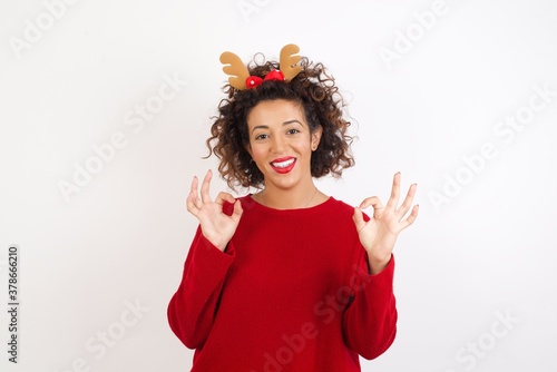 Young arab woman with curly hair wearing christmas headband showing both hands with fingers in OK sign. Approval or recommending concept