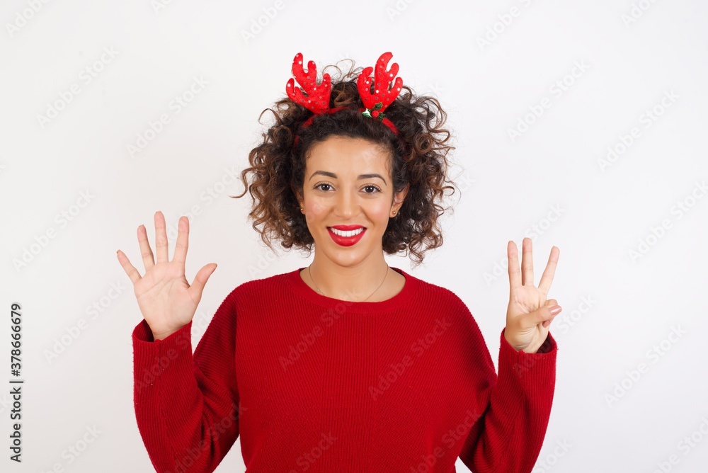 Young arab woman with curly hair wearing christmas headband standing on white background showing and pointing up with fingers number eight while smiling confident and happy. Young arab woman with curly hair wearing christmas headband standing on white background showing and pointing up with fingers number eight while smiling confident and happy.