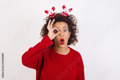 Young arab woman with curly hair wearing christmas headband standing on white background doing ok gesture shocked with surprised face, eye looking through fingers. Unbelieving expression.