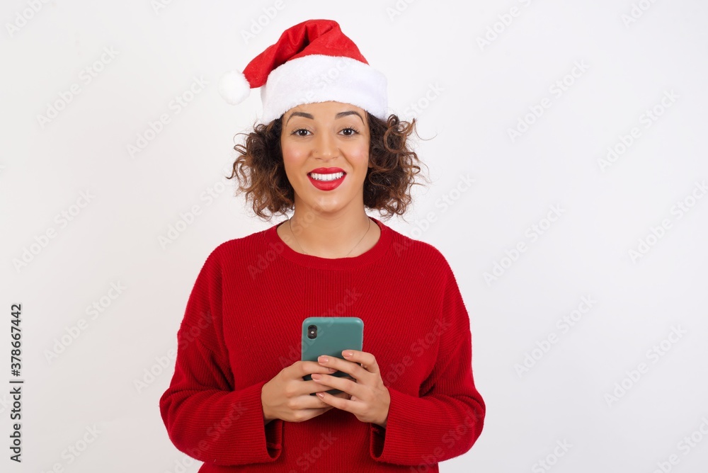 Young arab woman with curly hair wearing christmas hat on white background enjoys distant communication, uses mobile phone, surfs fast unlimited internet, has pleasant smile, makes shopping online, Young arab woman with curly hair wearing christmas hat on white background enjoys distant communication, uses mobile phone, surfs fast unlimited internet, has pleasant smile, makes shopping online,