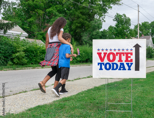 African American Mother walking to vote at Polling Station with Arm Around Mixed Race Son waving a Flag