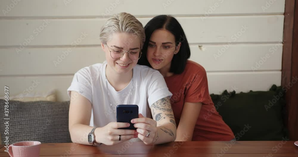 Brunette with loose hair hugs blonde girlfriend and couple talks looking into smartphone display against white wall slow motion Brunette with loose hair hugs blonde girlfriend and couple talks looking into smartphone display against white wall slow motion