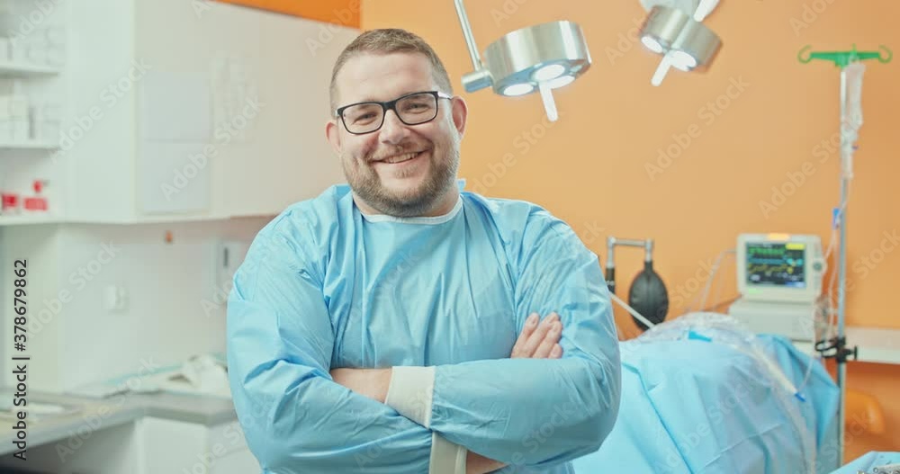 Male veterinarian, in glasses, in surgical outfit, crosses his arms over his chest and smiles, after the surgery operation on dog's leg. At the background - blurred orange walls, covered with surgical Male veterinarian, in glasses, in surgical outfit, crosses his arms over his chest and smiles, after the surgery operation on dog's leg. At the background - blurred orange walls, covered with surgical