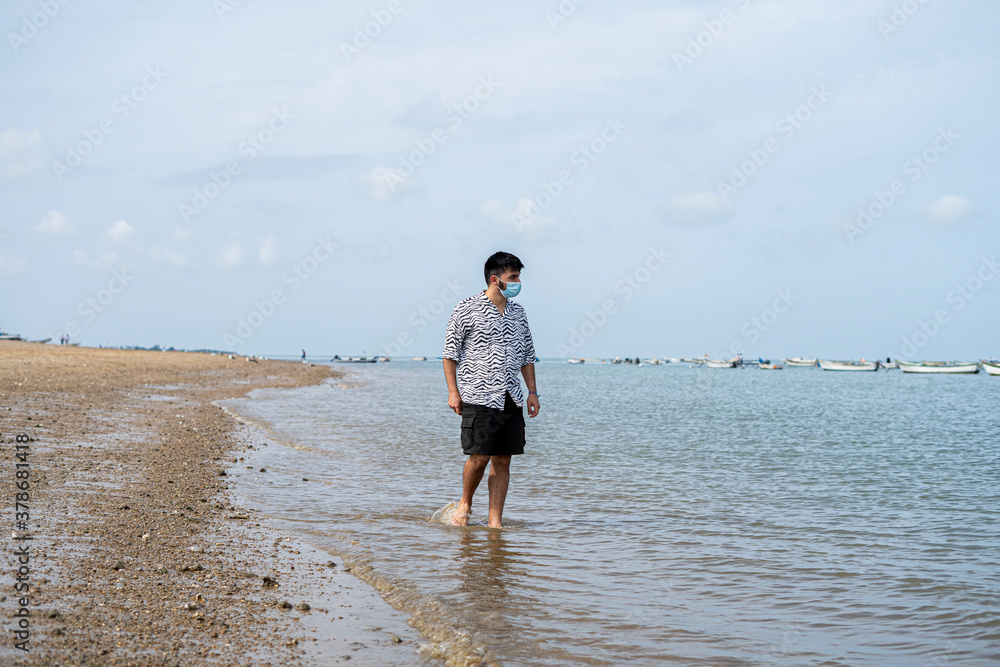 Hombre joven Adolescente con mascarilla caminando feliz por la orilla del mar en la playa Hombre joven Adolescente con mascarilla caminando feliz por la orilla del mar en la playa