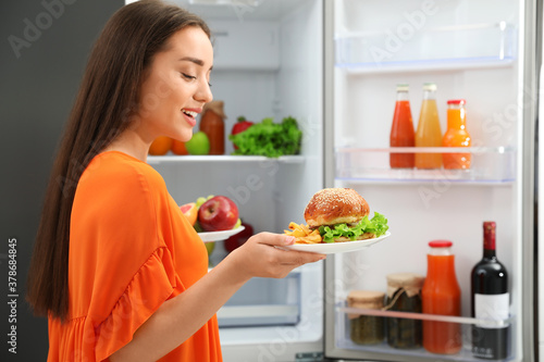 Woman choosing between fruits and burger with French fries near refrigerator in kitchen