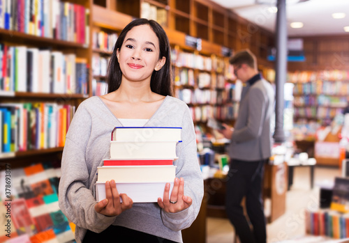 Happy young female student standing in library with pile of books in hands