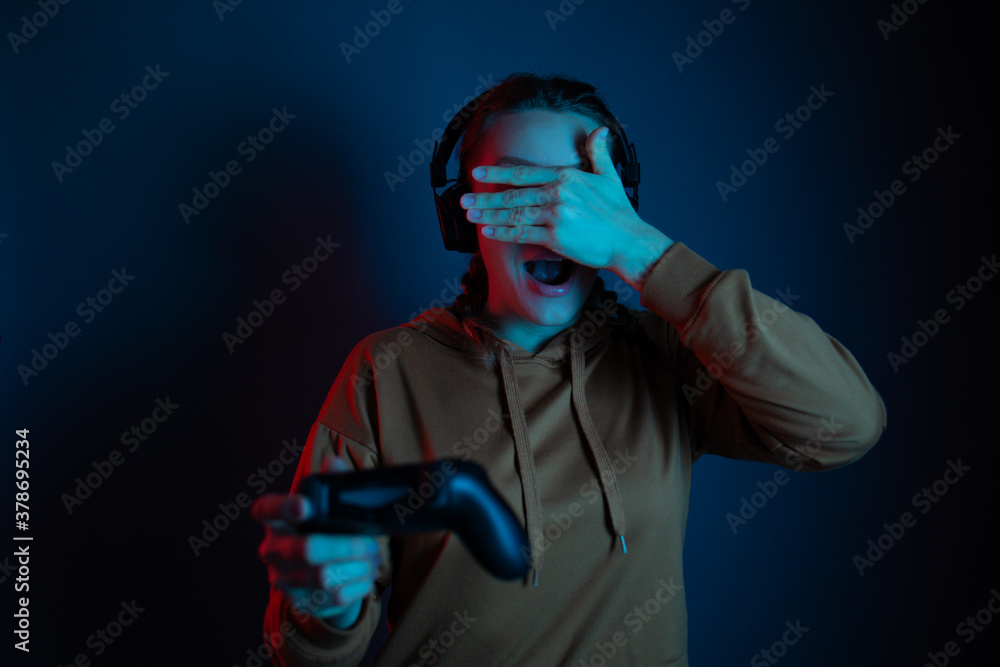 The gamer girl playing the online video game with joystick and headphones and close her eyes by the hand. Blue neon background. Gamer concept.