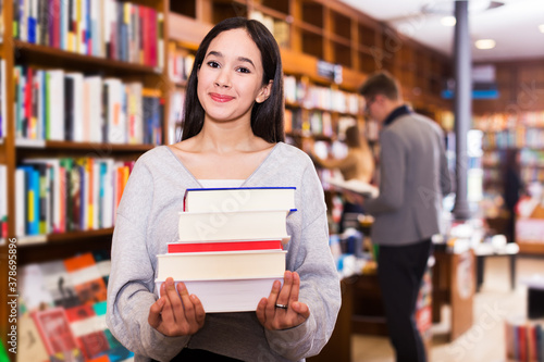Happy young female student standing in bookshop with pile of books in hands