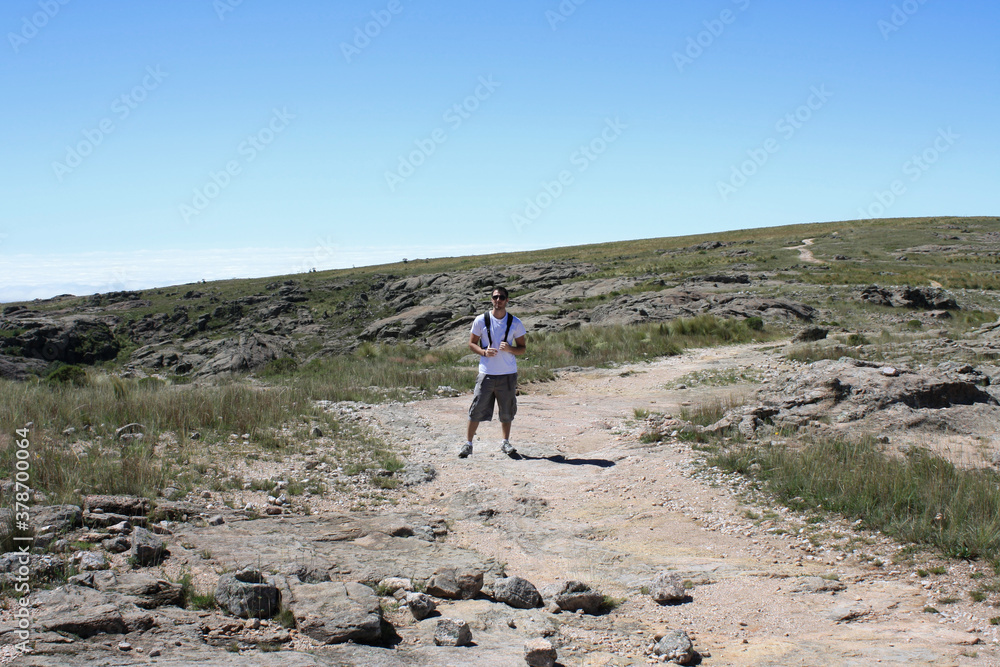 Hiker in the dirt path along the rocky mountaintop. Male young adult trekking the mountain summit. Beautiful view of the hills under a blue sky. 