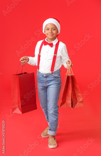 Cute African-American boy in Santa hat and with shopping bags on color background