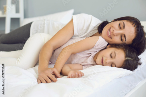 Happy relaxed mother and daughter cuddling on soft bed in quiet bedroom at weekend