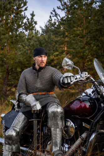 A medieval warrior in armor sits on a motorcycle with a sword in his hands on the background of the forest. Knight biker concept.