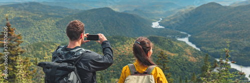 Hikers couple camping walking with backpacks in Quebec taking picture of view with phone in Autumn. Canada forest travel lifestyle banner. Tourists looking at Jacques Cartier National Park.