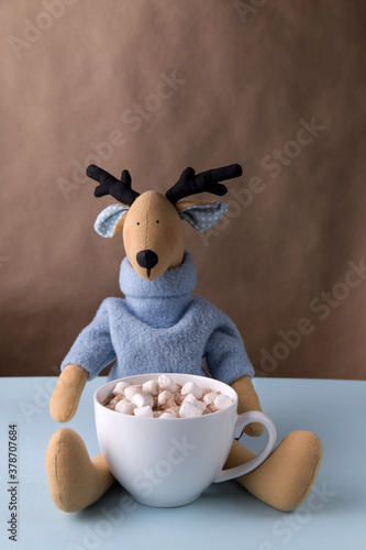 Christmas handmade reindeer toy sits on a brown background and holds a mug with cocoa or coffee decorated with marshmallows, vertical photo, minimalism