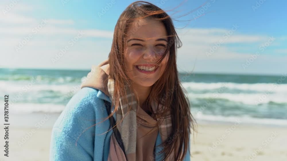 Young female with long hair flying on wind in a sky blue coat looks directly into the camera and laugh. Beach, fall, travel, harmony. Young female with long hair flying on wind in a sky blue coat looks directly into the camera and laugh. Beach, fall, travel, harmony.