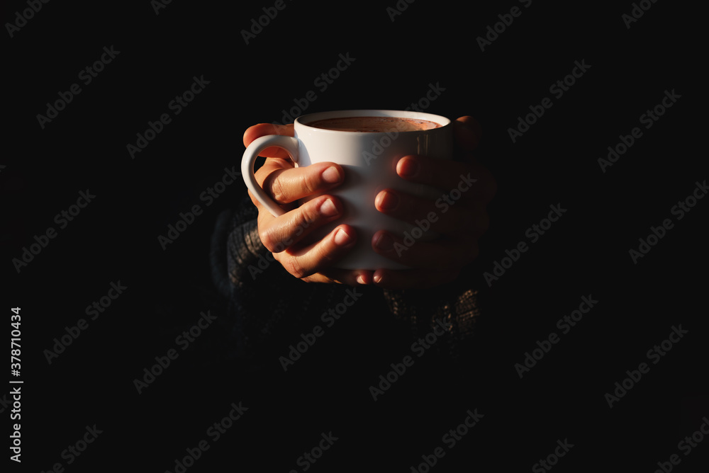 Female hands holding white cup of coffee on black background. Woman warming her hands on mug of hot beverage in the dark. Minimal style. Copy space. Low key