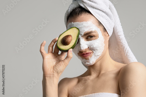 Close up studio portrait of funny girl in mask and white towel holding avocado isolated on gray background. Advertising of eco-friendly beauty products for healthy glowing skin. Morning beauty routine