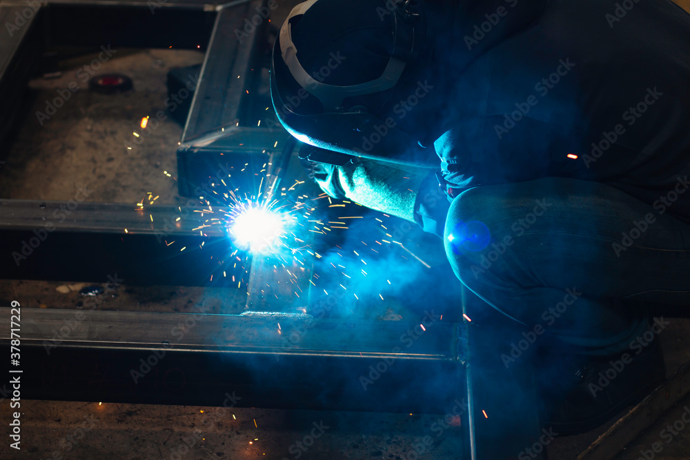 Welder is welding the steel in the factory. Weld the steel in dark. Workers wearing industrial uniforms and welding masks at a welding factory protect from welding sparks. Occupational safety concept Welder is welding the steel in the factory. Weld the steel in dark. Workers wearing industrial uniforms and welding masks at a welding factory protect from welding sparks. Occupational safety concept