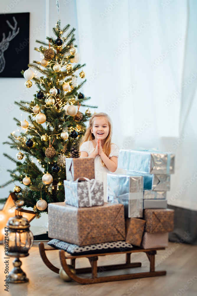a beautiful little girl in a white dress puts gifts on a sled near the Christmas tree. happy new year. retro-style. interior decoration for a holiday.