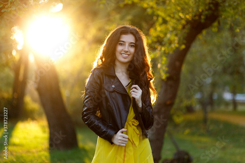 Fototapeta Naklejka Na Ścianę i Meble -  Cheerful girl with long hair, in a leather jacket and yellow dress in the park, looking at camera, against the background of the sunset.