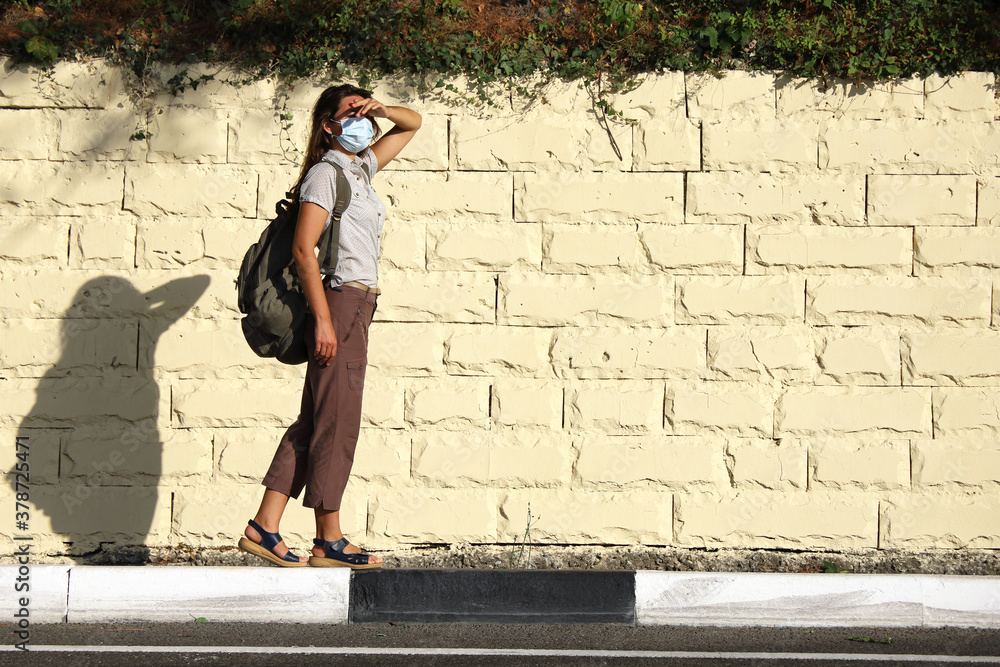 Young woman tourist, hitchhiker with backpack stands on the side of the road and waits for car or bus. She is wearing medical face mask and squinting in the bright sun, covering her eyes with her hand