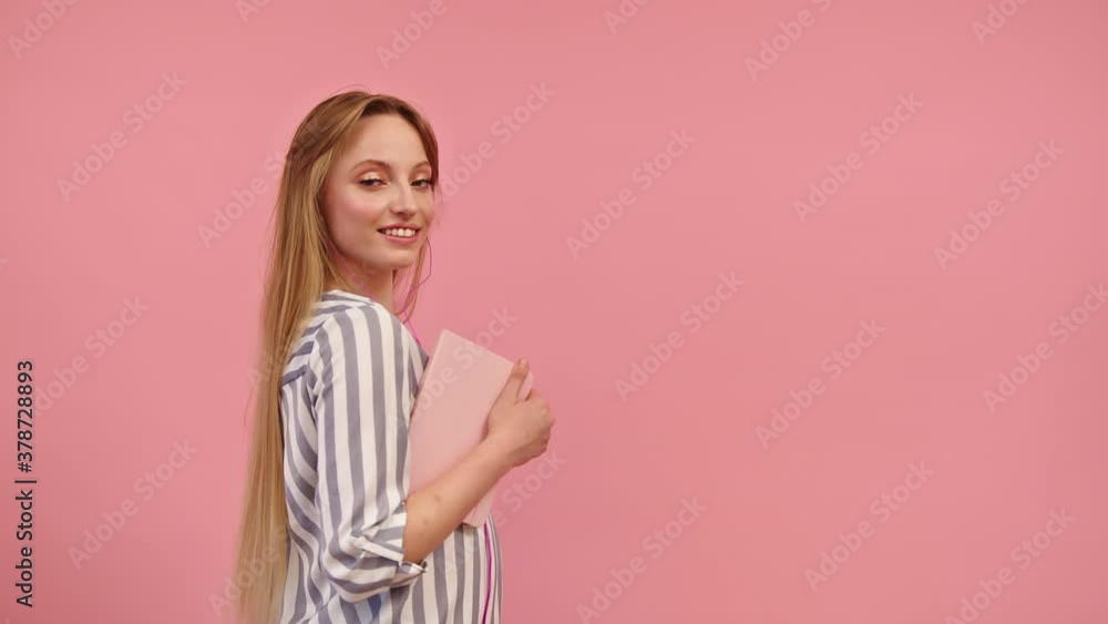 Attractive Woman Carrying A Pink Notebook On Her Arm Standing Sideways Against Pink Background - Half-Body Shot Attractive Woman Carrying A Pink Notebook On Her Arm Standing Sideways Against Pink Background - Half-Body Shot