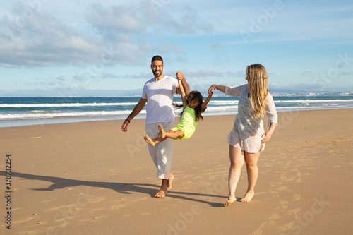 Joyful parents and kid having fun on beach, girl holding parents hands, jumping and hanging. Full length. Family outdoor activities concept