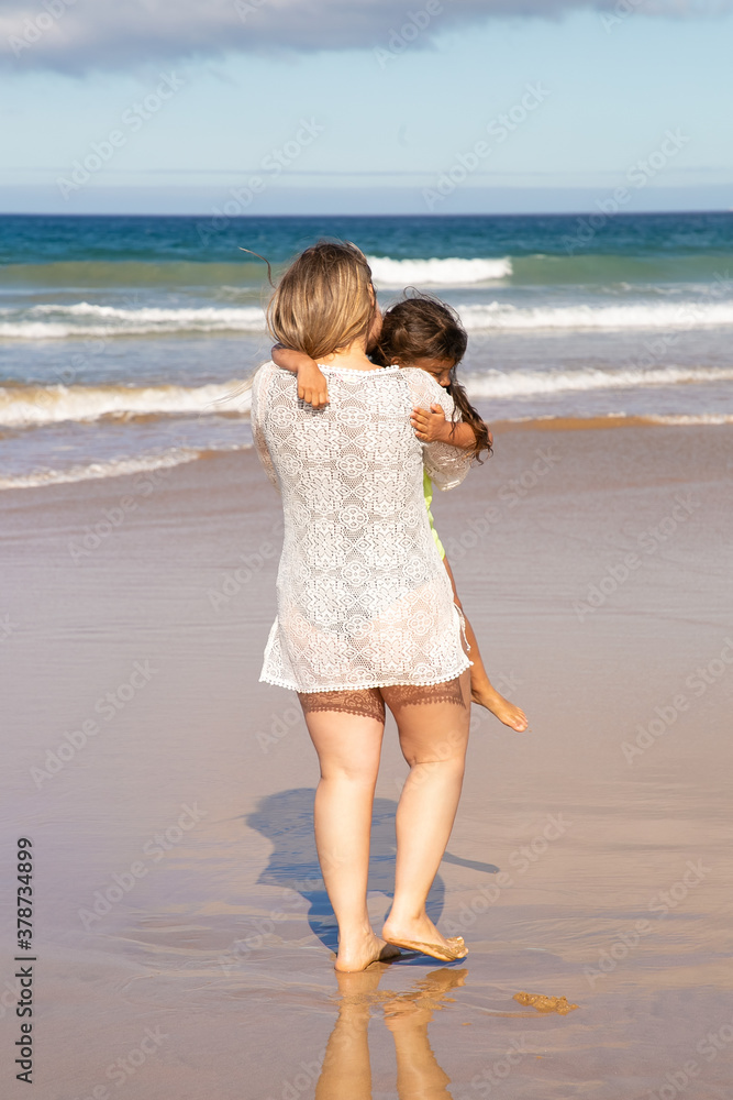Young mom spending leisure time with little daughter on beach at sea, holding kid in arms. Back view. Parenting and outdoor activities concept Young mom spending leisure time with little daughter on beach at sea, holding kid in arms. Back view. Parenting and outdoor activities concept
