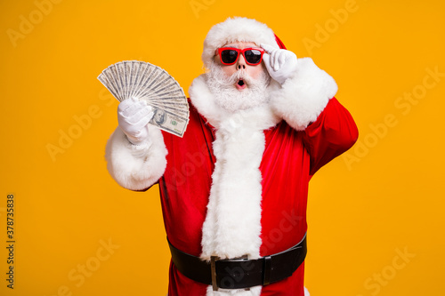 Portrait of his he nice attractive amazed astonished fat overweight bearded Santa holding in hand usd cash 100 hundred salary deposit isolated bright vivid shine vibrant yellow color background © deagreez