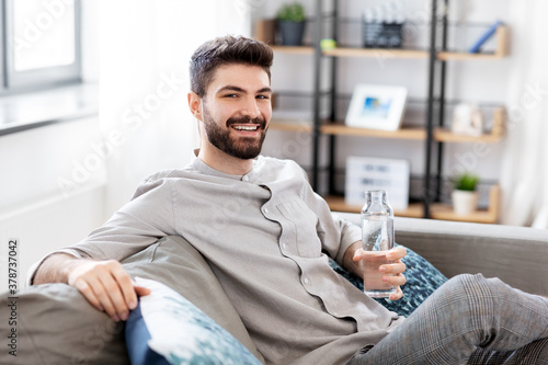 people, sustainability and leisure concept - happy smiling young man sitting on sofa and drinking water from glass bottle at home