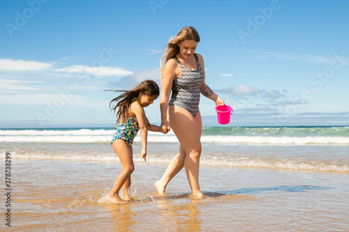 Mom and little girl walking ankle deep in sea water and wet sand, picking shells into bucket. Bright blue sea with waves and white foam in background. Family summer holidays concept
