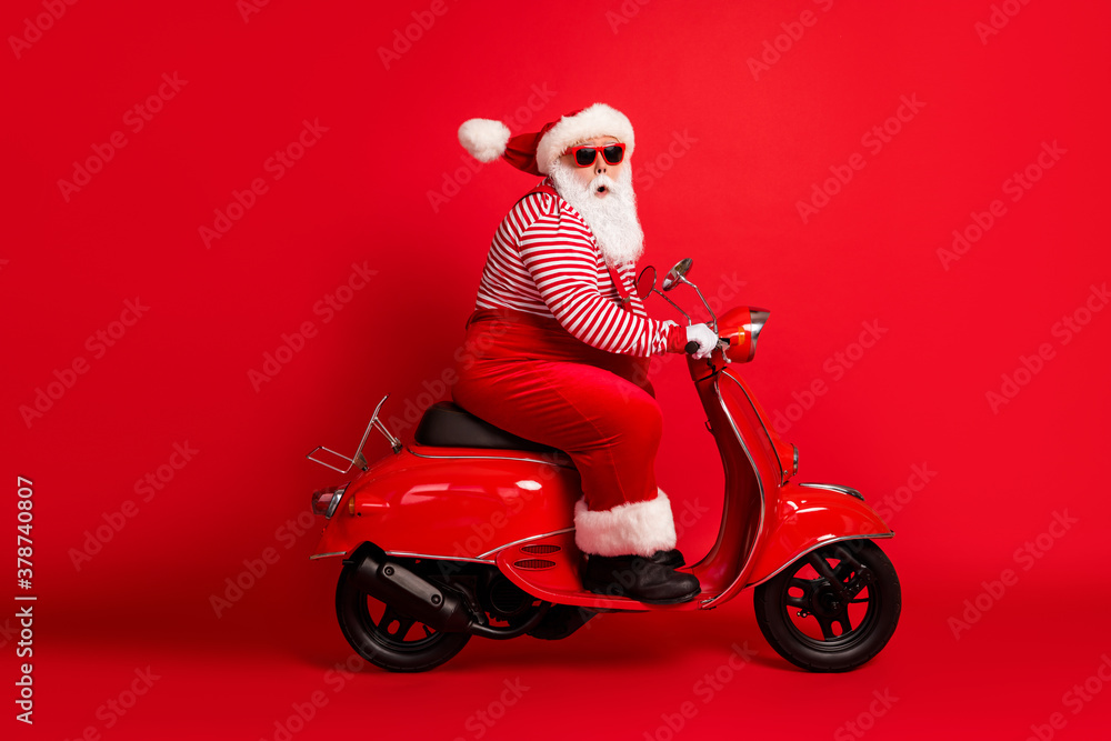 Full length profile photo of excited grandfather grey beard ride retro moped look camera wear santa claus x-mas costume suspenders sunglass striped shirt cap isolated red color background Full length profile photo of excited grandfather grey beard ride retro moped look camera wear santa claus x-mas costume suspenders sunglass striped shirt cap isolated red color background